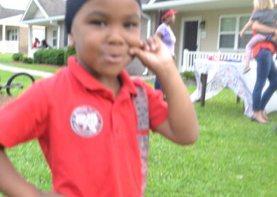 An African-American child grins while grabbing his cheek and putting his hand on his hip. He is wearing a red shirt and having fun outside.