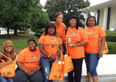 A group of African American women and girls in orange shirts smile outside of a government building. Their shirts read "Raise the Age: North Carolina Youth Deserve the Same Opportunities!"
